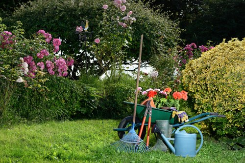 A well-maintained garden scene featuring a lush, dense green lawn with a mix of flowering shrubs and bushes, including pink and purple azaleas and yellow-green foliage in the background. The garden is bordered by a variety of plants and flowers, creating a vibrant and colourful atmosphere. In the foreground, gardening tools such as a blue rake, a garden fork, a watering can, and a large waste bin are arranged on the grass, indicating ongoing or recent gardening activities. The environment appears to be in natural daylight with gentle shadows, suggesting a mild weather day, and the garden setting is typical of outdoor residential spaces found in Mill Hill, North London. The scene reflects a tidy and active outdoor space suitable for gardening services from Landscapers Mill Hill, capturing the natural features and plant varieties common in the area, with the focus on outdoor maintenance and garden care.