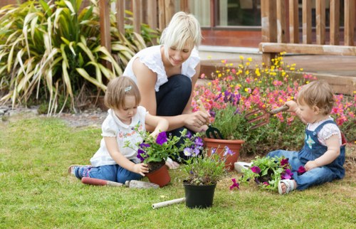 A woman and two young children are working together in a garden on a clear day, planting and tending to potted flowers. The outdoor space features a well-maintained lawn with vibrant green grass, bordered by a wooden deck or patio area in the background. The garden includes a variety of flowering plants in shades of pink, purple, and yellow, arranged in flower beds and pots, creating a colorful, natural landscape. The children are seated on the grass, each handling flower pots filled with blooming plants, while the woman assists and supervises, smiling softly. Surrounding the garden are tall, ornamental grasses and shrubs, adding texture and structure to the scene. The environment appears sunny with soft natural light, indicating favorable weather for outdoor gardening activities, supporting natural connection to gardening and landscaping services offered by Landscapers Mill Hill, as seen in the context of Mill Hill in London, UK.