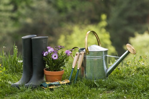 A collection of gardening tools and accessories arranged on a lush green lawn in a backyard, including a pair of tall black rubber boots, a small terracotta pot filled with purple daisies, a silver metal watering can with a wooden handle, and a set of hand tools with light-colored wooden handles. The background displays a softly blurred garden scene with trees and foliage, illustrating a well-maintained outdoor space suitable for gardening activities. The natural lighting suggests a bright, possibly overcast day, emphasizing the fresh green tones of the grass and the vibrant colours of the flowers and tools, aligning with gardening and landscaping services provided by Landscapers Mill Hill in the Mill Hill area near London, and reflecting a typical suburban garden setup in the UK.