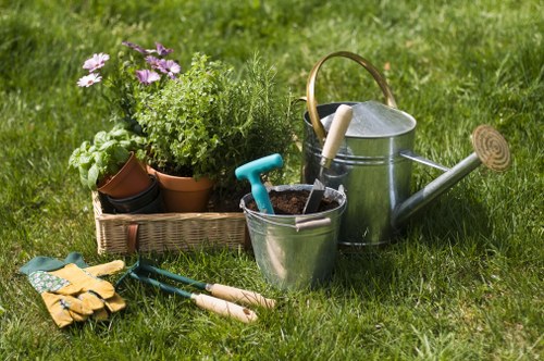 A tidy garden scene in a residential outdoor space features a lush green lawn with well-maintained grass. In the foreground, there are gardening tools including a pair of yellow gardening gloves, a small hand rake, and a trowel placed on the grass. To the left, a woven basket holds a variety of potted plants, including flowering plants with purple blossoms and green foliage, arranged on the lawn. Behind the plants, a metal watering can with a long spout and wooden handle is positioned, ready for watering tasks. Nearby, a metal bucket contains soil or compost, with a gardening trowel placed inside it. The background reveals a well-kept hedge or shrubbery, providing a natural boundary to the garden, while further elements like paved areas or flower beds are not visible. The scene appears to be set during daylight with natural light, indicating fair weather, which supports outdoor gardening activities typical of a residential garden in the Mill Hill area.