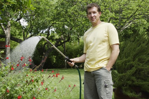A man wearing a pale yellow t-shirt and grey cargo shorts stands in a lush, well-maintained garden, holding a garden hose that is spraying water over a flower bed with red blossoming flowers. The garden features a neatly cut lawn in the foreground, bordered by vibrant green shrubs and trees with dense foliage in the background. Behind him, there are tall, leafy trees providing shade, and a hedge creating a natural boundary along the garden's edge. The scene is set outdoors on a clear day with natural sunlight illuminating the garden, emphasizing the healthy, thriving plants and the overall landscaped environment. This setting demonstrates typical features of a landscaped private garden, suitable for professional gardening and outdoor maintenance services by companies like Landscapers Mill Hill, located near Mill Hill and the N20 postcode area, with the garden designed to support lush plant growth and outdoor enjoyment.