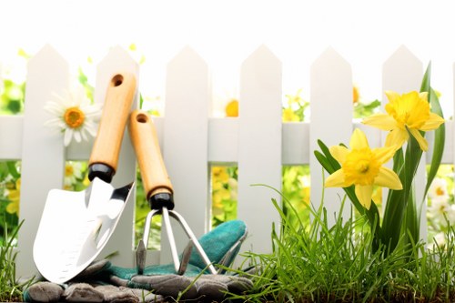 The image depicts a garden scene in front of a white picket fence, featuring a collection of gardening tools including a small trowel, a hand cultivator, and a spade, all with wooden handles and metal working parts. These tools are placed on the ground near some lush green grass and a few bright yellow daffodil flowers with long green leaves growing beside them. In the background, the fence is set against a blurred garden with hints of additional greenery and flower beds, suggesting a well-maintained outdoor space typical of a residential garden in Mill Hill, London. The lighting indicates a clear day with natural sunlight, enhancing the vivid colors of the flowers and foliage. This arrangement reflects typical gardening activities such as planting, weeding, or preparing the soil for upcoming garden work, aligning with services offered by companies like Landscapers Mill Hill specializing in lawn care, plant maintenance, and outdoor improvements in the local area. The overall scene communicates a tidy, inviting garden environment ready for ongoing horticultural care, fitting seamlessly into a landscape maintenance or gardening service context for Mill Hill residents.