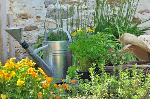 A lush garden scene featuring a variety of vibrant flowering plants and greenery, with bright yellow and orange flowers in the foreground. Behind the flowers, there is an assortment of potted plants, including leafy green foliage. To the left, a galvanized metal watering can sits next to a garden trowel, both placed among the plants. The background includes a rustic stone wall, with taller grasses or reeds growing nearby, adding natural vertical elements. The environment appears well-maintained, reflecting typical outdoor landscaping found in Mill Hill, London, with natural light illuminating the garden in a daytime setting. The arrangement suggests a home garden area with a focus on horticultural care and outdoor maintenance, aligning with gardening services provided by Landscapers Mill Hill.