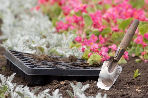 A gardening scene in a landscaped garden with freshly turned soil, a black plastic seed tray placed on the ground, and a small garden trowel with a wooden handle partially embedded in the soil. In the background, there are vibrant pink and white flowering plants, likely petunias or similar annuals, alongside neatly arranged green foliage. The garden area features a mix of flower beds and soil patches, with natural daylight illuminating the scene and a slightly blurred background that suggests a well-maintained outdoor space in Mill Hill, London, consistent with local gardening and landscaping services provided by Landscapers Mill Hill.