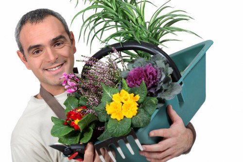The image depicts a smiling man in casual clothing holding a garden watering can filled with a variety of colorful flowering plants, including yellow, purple, pink, and red blooms, with lush green leaves. Behind him, a small section of a well-maintained garden is visible, featuring a neatly trimmed hedge and some trees in the background, suggesting an outdoor space typical of residential gardens in Mill Hill. The scene appears to be outdoors during daylight, with natural lighting highlighting the vibrant plant colors and the greenery of the garden environment. This setup reflects typical gardening and plant care activities that a professional landscaper or gardener, such as Landscapers Mill Hill, might undertake to enhance outdoor spaces and maintain garden aesthetics, including plant planting, flowering, and general outdoor plant management.
