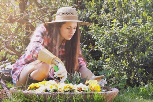 A young woman wearing a wide-brimmed straw hat, plaid shirt, and gardening gloves is kneeling on a lush green lawn in a residential garden, tending to a colourful flower bed. The flower bed is edged with a low brick boundary and contains a variety of vibrant yellow and white flowers. Behind her, dense foliage and tall bushes create a natural backdrop, with sunlight filtering through the leaves, indicating a clear, sunny day. To her right, a section of neatly mowed grass leads towards a background of mature trees and shrubs, providing shade and privacy. The garden appears well-maintained, with a mix of lawn, soil borders, and flowering plants, suggesting professional outdoor care typical of local landscaping services in Mill Hill. The scene captures a peaceful moment of gardening activity, emphasizing the natural textures of the grass, soil, and flowering plants, suitable for a landscaping or gardening service webpage focused on flower bed creation and outdoor garden maintenance in the Mill Hill area of London.