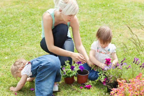 A woman with short blonde hair, dressed in a light blue sleeveless top and dark blue jeans, is kneeling on a well-maintained lawn in a garden area with vibrant flower beds. She is engaged in planting or tending to potted plants, with her right hand reaching into one of the pots. Beside her, two young children, a girl with long light brown hair in a white top with pink floral accents, and a boy with short blonde hair in a blue shirt and jeans, are actively involved in garden activities; the girl is handling soil or flowers, while the boy is kneeling close to the ground, seemingly exploring or planting. The garden features a lush green lawn, colorful flower beds with pink, purple, and orange blooms, and a clear outdoor environment with bright natural lighting suggesting a sunny day. The scene exemplifies outdoor gardening activities suitable for family involvement, reflecting the services offered by Landscapers Mill Hill's gardening division in the local area near Mill Hill, London, with a focus on maintaining attractive front or back gardens. The overall setting emphasizes healthy plant growth, natural textures of soil and flowering plants, and a tidy, inviting outdoor space.