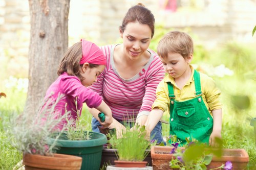 In this outdoor garden scene, a woman is engaged in planting activities with two young children, a girl and a boy, in a lush, well-maintained backyard. The garden features a variety of potted plants, including herbs and flowering plants, arranged on a surface near a mature tree with textured bark and leafy branches providing dappled shade. The children are dressed in colorful clothing, with the girl wearing a pink headscarf and the boy in a yellow shirt with green overalls, both actively participating in gardening tasks such as planting or watering. The background displays a neatly tended lawn, bordered by a hedge or shrubbery, with soft natural light suggesting a mild, sunny day. The overall environment reflects a tidy, landscaped outdoor space suitable for gardening activities with natural tones of green, brown, and vibrant plant colours, contributing to a peaceful and inviting atmosphere typical of residential gardens in Mill Hill that professional gardeners like Landscapers Mill Hill might maintain. The scene emphasizes nurturing outdoor environments through planting and care, connecting visually with gardening services aimed at enhancing local outdoor spaces.